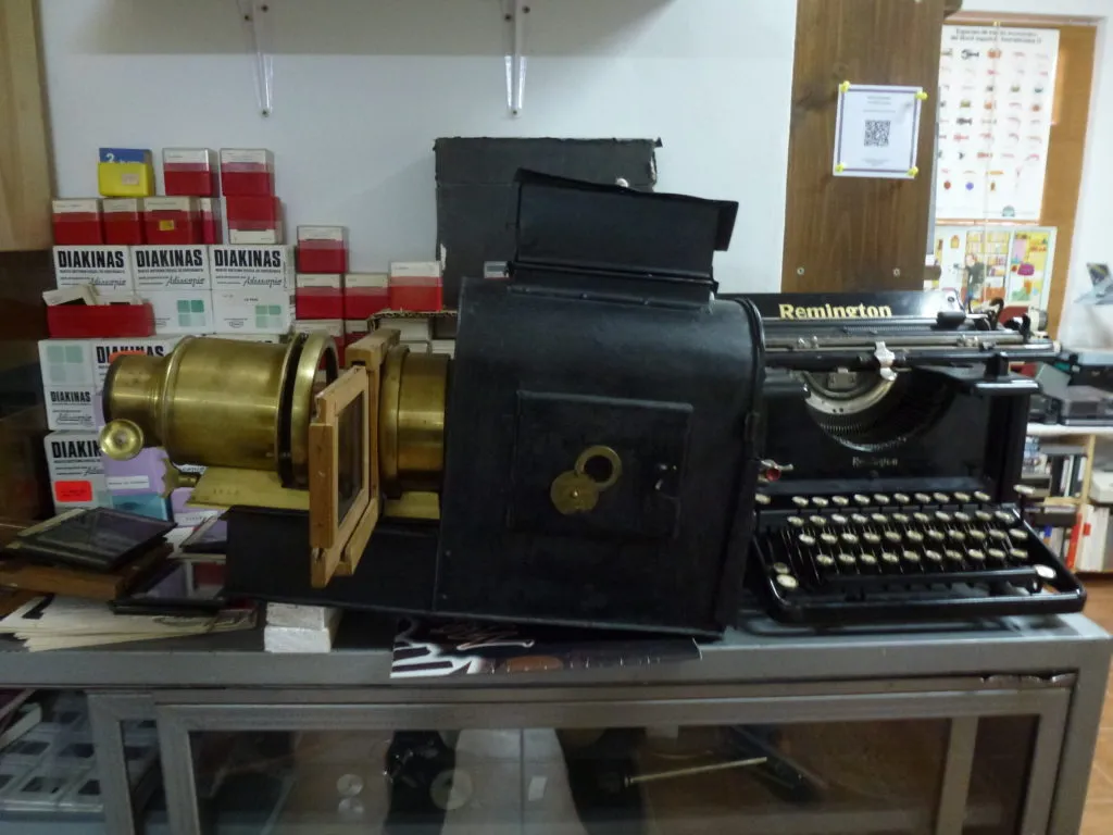 Vista de una mesa de trabajo en el museo con una máquina de escribir negra de época situada a la derecha y un proyector óptico antiguo con cuerpo negro y lentes de latón en el centro y la izquierda.