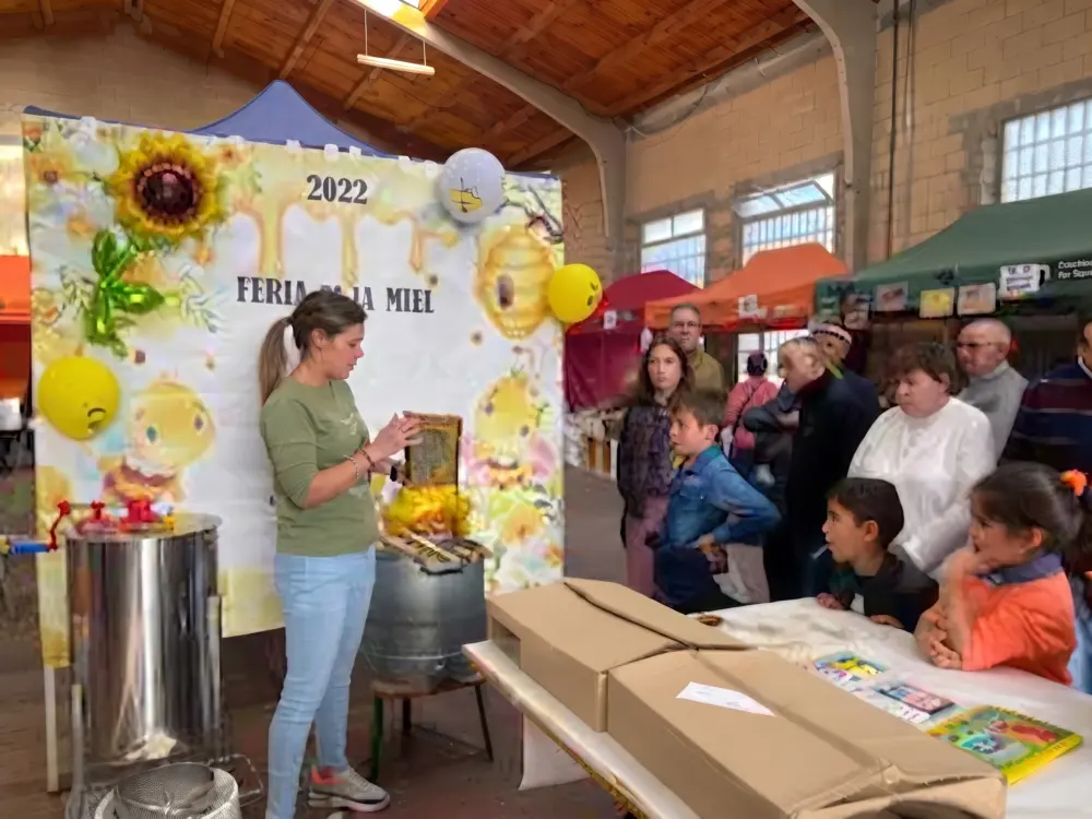 Taller o demostración sobre la miel en una feria rural, con una monitora mostrando un panal a un grupo de asistentes y niños.
