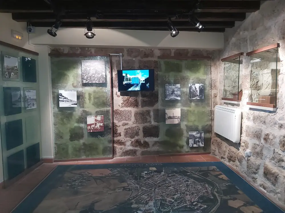 Sala museística con muro de piedra, fotografías históricas en paneles de vidrio, monitor multimedia y plano aéreo en el suelo de Medina de Rioseco.