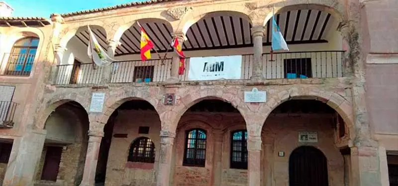Comparativa visual deslizante de la fachada de un edificio histórico porticado con soportales de piedra en la planta baja y un balcón corrido con arcos y banderas en la planta superior.