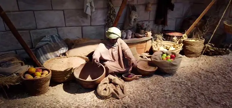 Maniquí sentado de una persona con túnica y turbante clasificando alimentos en una recreación histórica con paredes de piedra y cestas de mimbre llenas de fruta.