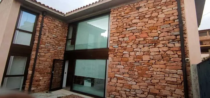 Fachada exterior de un edificio moderno revestido en piedra rústica y grandes ventanales de cristal con marcos oscuros, incluyendo una puerta de entrada lateral.