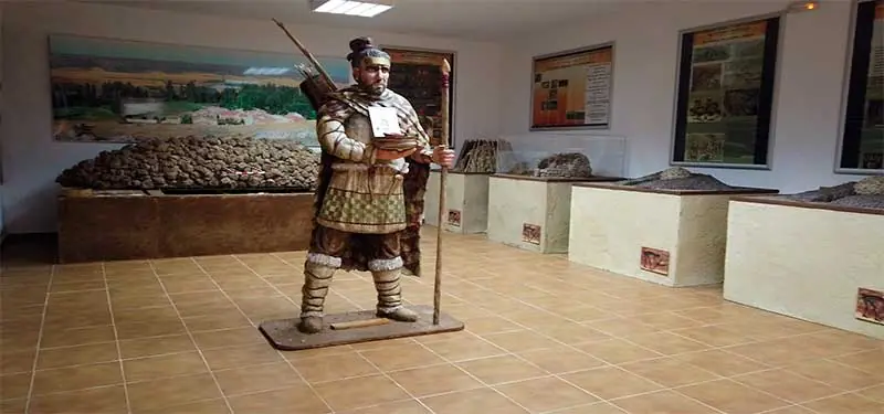Maniquí a tamaño real de un guerrero antiguo con lanza y arco en el interior de una sala de exposición con maquetas arqueológicas y paneles informativos.