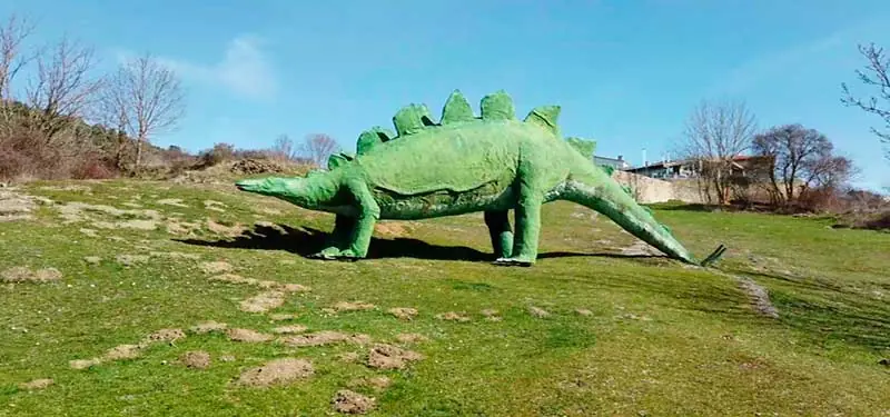 Escultura a tamaño real de un dinosaurio estegosaurio de color verde situada en una pradera al aire libre.