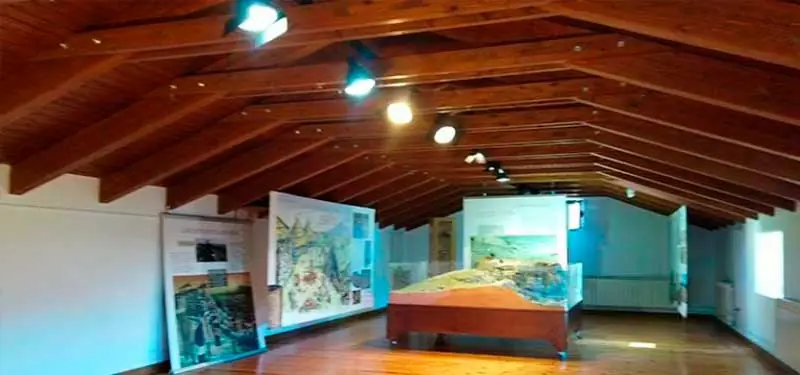 Sala de museo abuhardillada con suelo de madera, techo con vigas de madera a dos aguas, paneles informativos laterales y una gran maqueta central sobre soporte de madera.