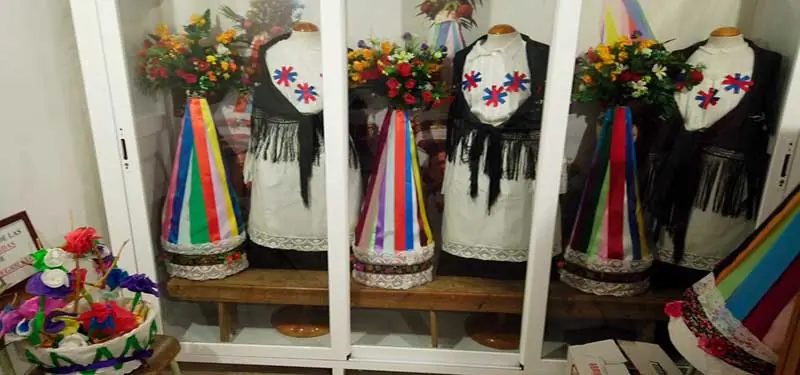 Vitrina de museo que exhibe tres maniquíes con trajes tradicionales regionales blancos y negros, acompañados de grandes tocados cónicos decorados con cintas de colores y arreglos florales.