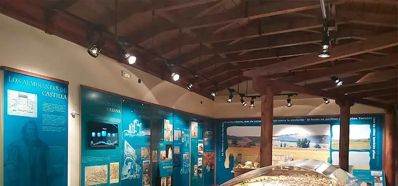 Un banner horizontal que muestra el interior de una sala de exposición con un rústico techo de vigas de madera y paredes de color azul petróleo con paneles de exhibición iluminados. En la parte inferior, hay una superposición blanca con el texto parcial "centro-".
