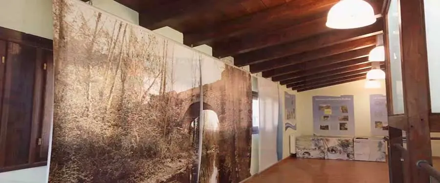 Vista interior de un pasillo de museo con un gran mural fotográfico de un puente antiguo y arbolado a la izquierda, y paneles informativos en la pared del fondo bajo un techo de vigas de madera.