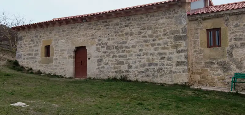 Fachada lateral de un edificio tradicional de piedra con una puerta y ventanas de madera, situado sobre un terreno de césped.