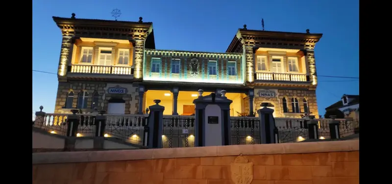 Fachada exterior de un edificio histórico de dos plantas con torres laterales, piedra en las esquinas y un letrero que dice "NIÑOS", rodeado por un muro bajo y un cielo azul despejado.