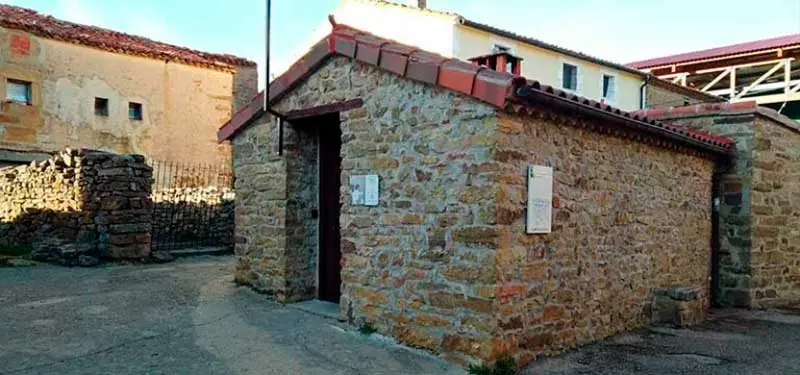 Exterior de un pequeño edificio rústico de piedra con tejado a dos aguas y puerta central, situado en una plaza empedrada.