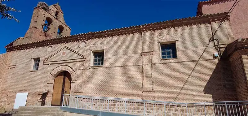 Una toma exterior lateral de una iglesia rústica de ladrillo española bajo un cielo azul despejado. La fachada texturizada cuenta con un gran portal de arco de medio punto rematado por un frontón triangular ciego. Arriba hay dos ventanas rectangulares enrejadas con marcos blancos. En el extremo izquierdo se eleva una espadaña de tres arcos vacíos para campanas, con un remate decorativo.