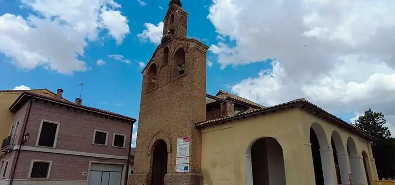 Fotografía exterior de una iglesia tradicional de ladrillo con una alta espadaña central y un pórtico lateral de arcos, ubicada junto a un edificio residencial rojizo bajo un cielo con nubes blancas.