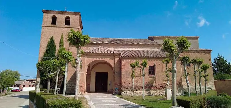 Fotografía horizontal de la fachada de una iglesia rústica de ladrillo con un campanario cuadrado y un pórtico porticado de tres arcos. Varios árboles podados están plantados frente a ella, bajo un cielo azul despejado con nubes dispersas.