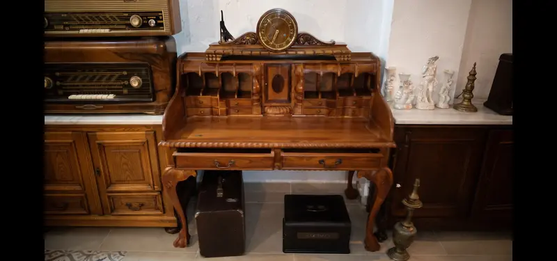 Antiguo escritorio de madera con persiana, compartimentos superiores y un reloj de sobremesa, flanqueado por muebles de madera y radios antiguas.