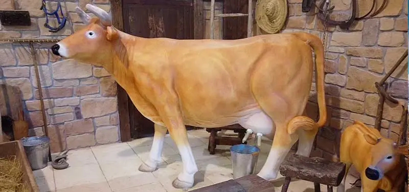 Escultura a tamaño real de una vaca de color marrón claro y blanco en el interior de un establo rústico con paredes de piedra.