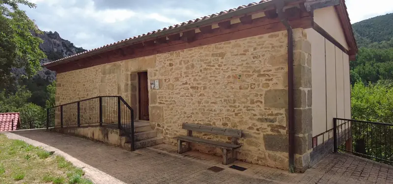 Edificio rural de piedra y madera con rampa de acceso y un banco de madera en la fachada, rodeado de naturaleza.