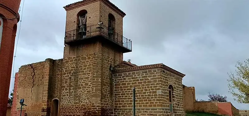 Fotografía a nivel de calle de una torre de iglesia histórica de piedra antigua con un balcón mirador de hierro forjado y campanas bajo un cielo nublado.