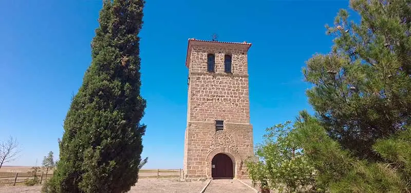 Torre de piedra antigua de una iglesia con un ciprés a la izquierda y un árbol frondoso a la derecha bajo un cielo azul brillante.