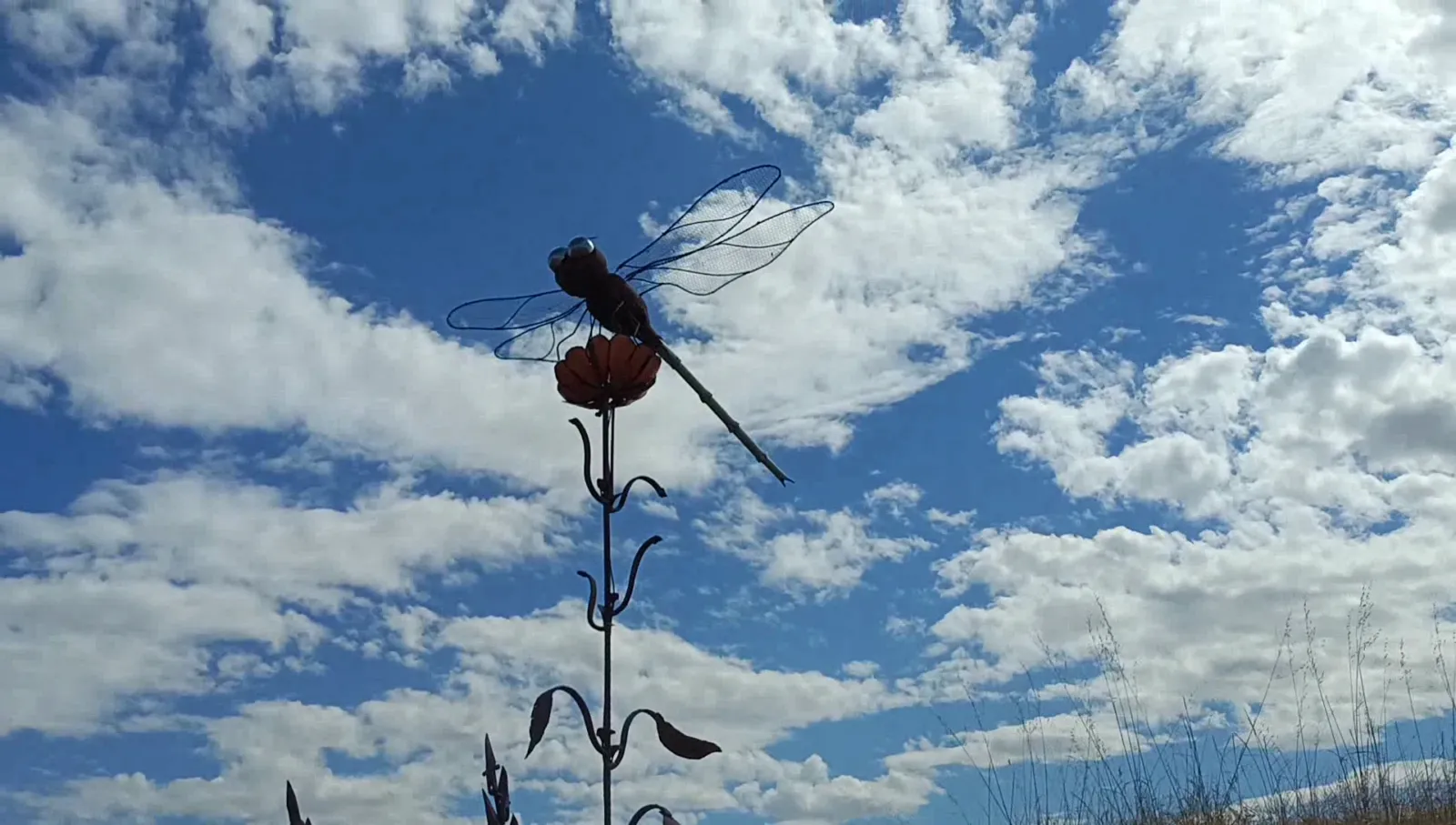 Escultura de metal forjado con forma de libélula posada sobre una flor, destacando la silueta de sus alas contra un cielo azul claro lleno de nubes blancas dispersas. En la esquina inferior derecha se aprecia un poco de hierba seca alta.